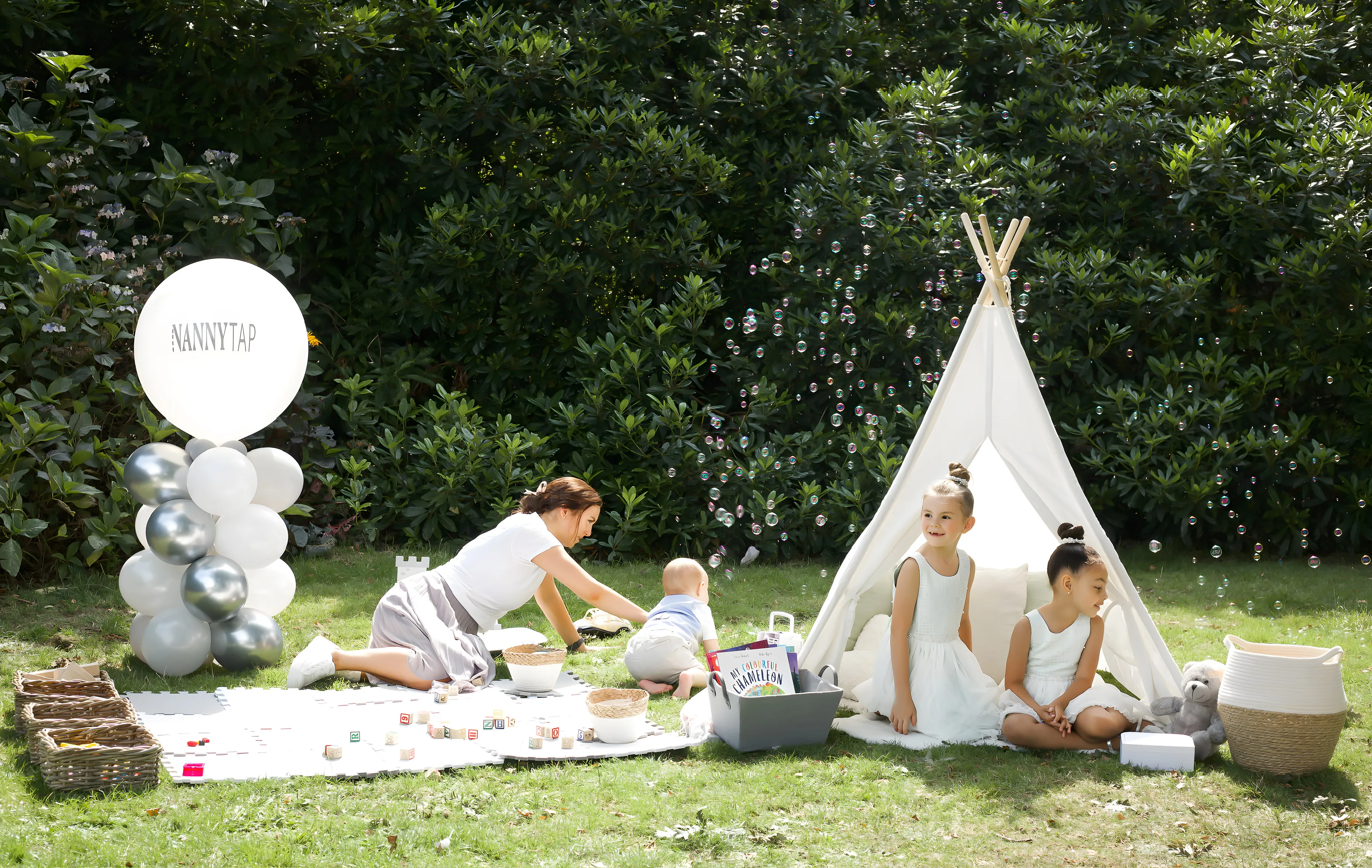The full NannyTap crèche staged on a lawn: a giant white NannyTap balloon on a column of silver and white balloons, a nanny kneeling on a foam play mat with a baby and wooden blocks, baskets of toys, bubbles drifting past, and a white teepee where two flower girls sit.